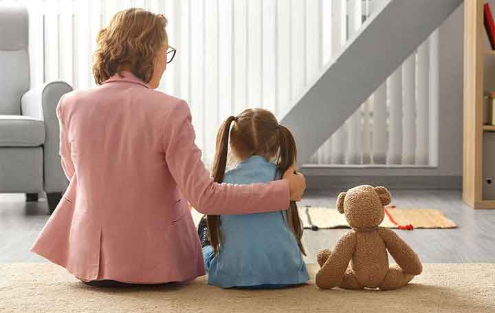 Imagen de una niña, su mamá y su oso sentados de espalda en una alfombra platicando